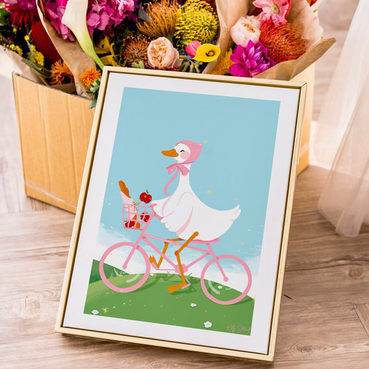 Framed illustration of a cartoon duck riding a pink bicycle with flowers in the background.