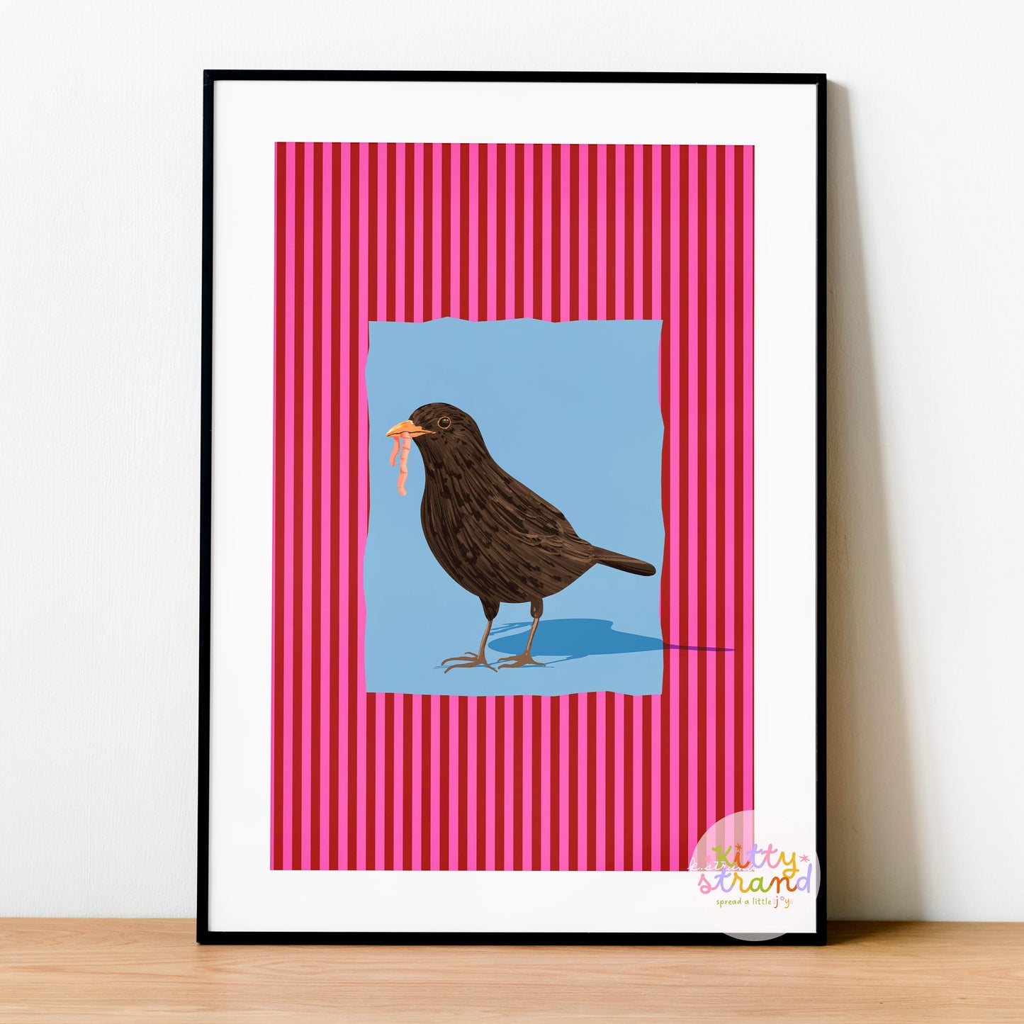 pink and blue bird-inspired wall art. Wall art with a vibrant striped pink background and a soft blue geometric square sits behind an original hand illustration of a blackbird with a worm in its mouth. Colourful, bold dopamine decor art print for nature lovers displayed in a wood frame