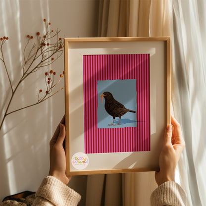 pink and blue bird-inspired wall art. Wall art with a vibrant striped pink background and a soft blue geometric square sits behind an original hand illustration of a blackbird with a worm in its mouth. Colourful, bold dopamine decor art print for nature lovers displayed in a wood frame