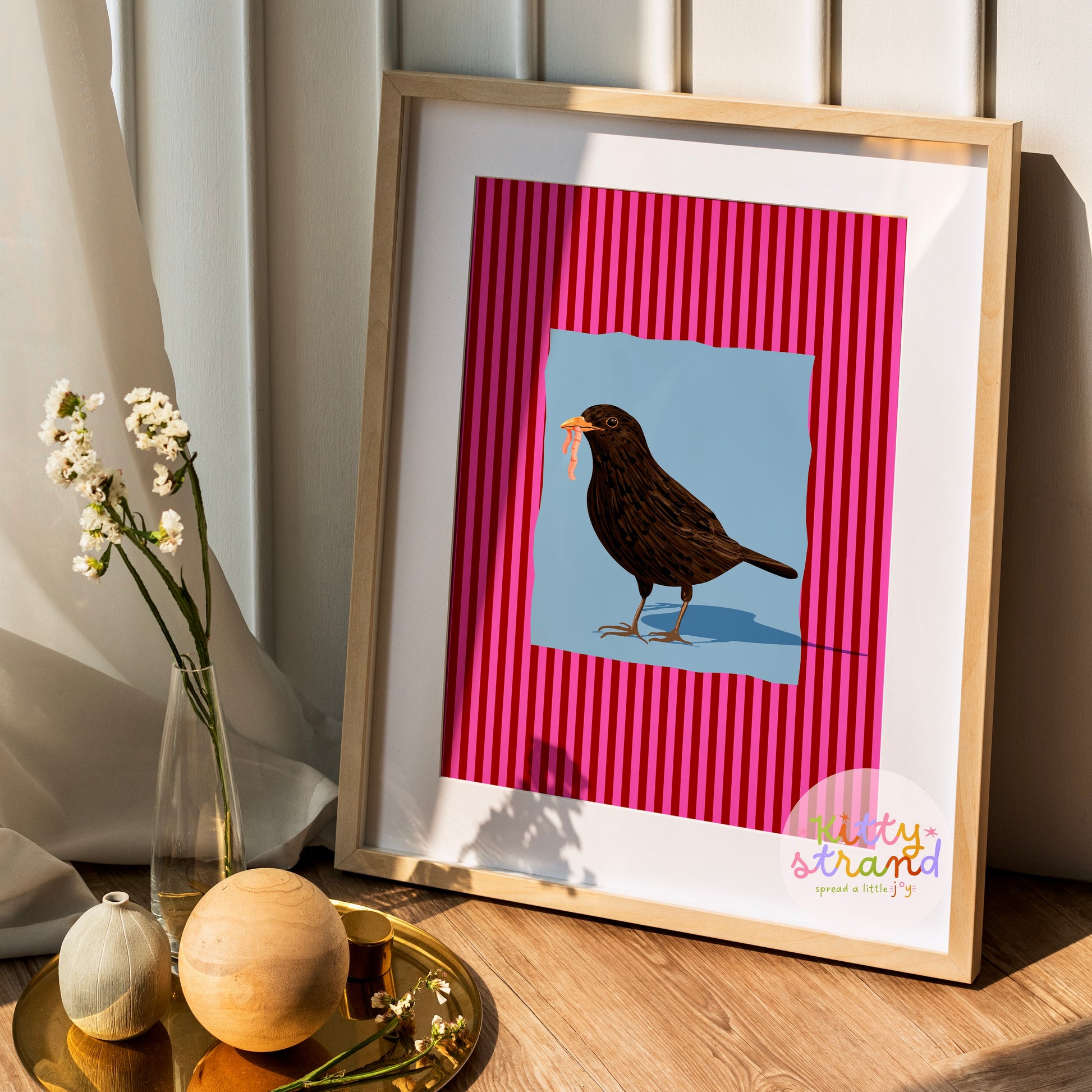 pink and blue bird-inspired wall art. Wall art with a vibrant striped pink background and a soft blue geometric square sits behind an original hand illustration of a blackbird with a worm in its mouth. Colourful, bold dopamine decor art print for nature lovers displayed in a wood frame
