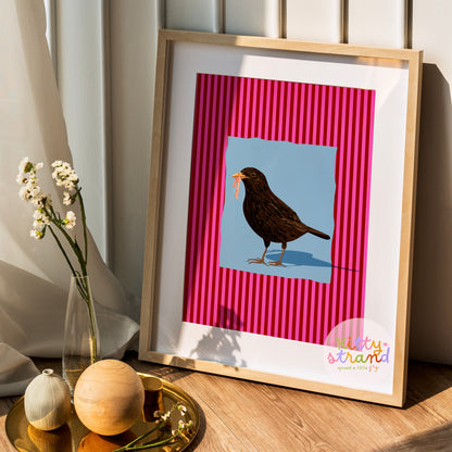 pink and blue bird-inspired wall art. Wall art with a vibrant striped pink background and a soft blue geometric square sits behind an original hand illustration of a blackbird with a worm in its mouth. Colourful, bold dopamine decor art print for nature lovers displayed in a wood frame