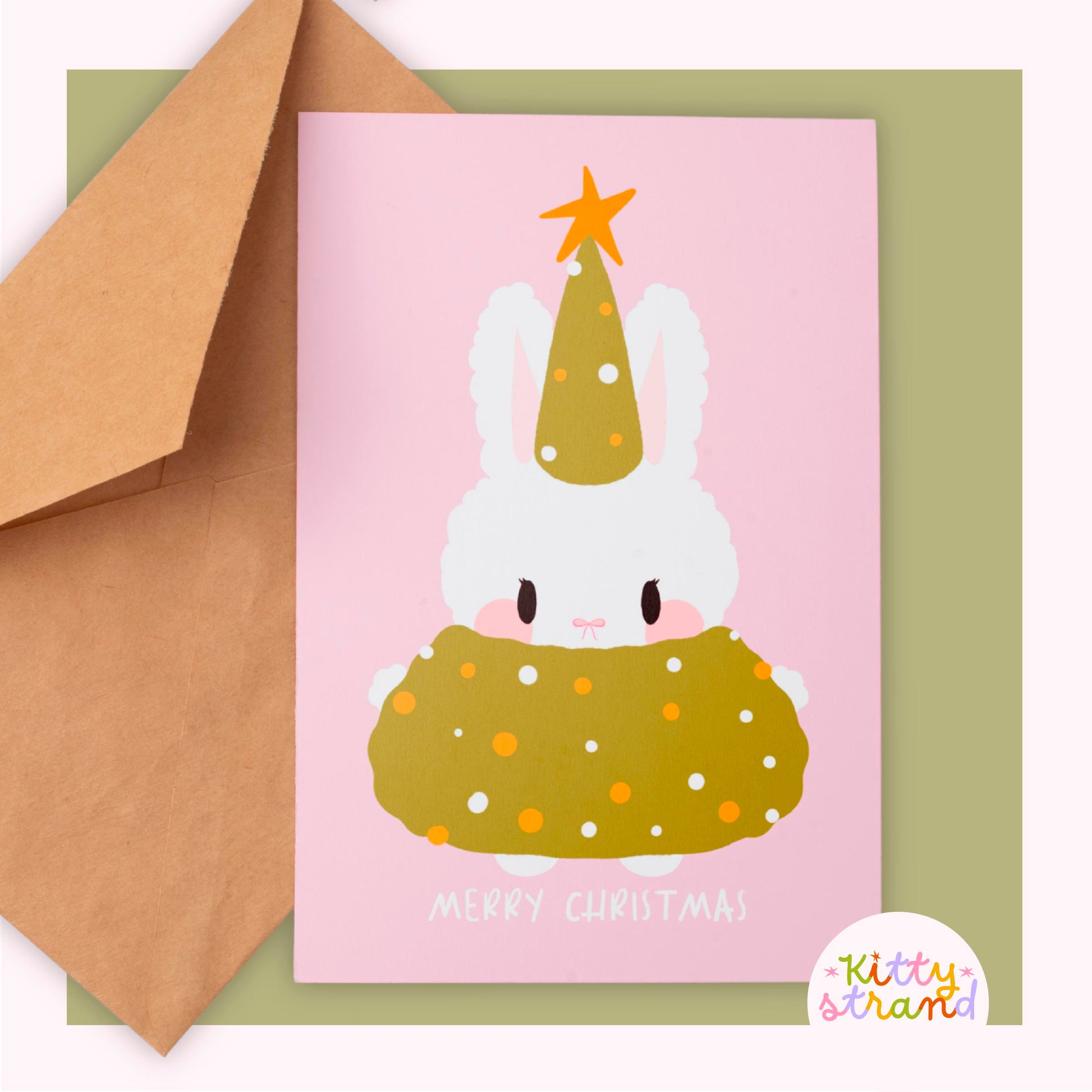Christmas card with a bunny wearing a green hat and sweater, next to a brown envelope.
