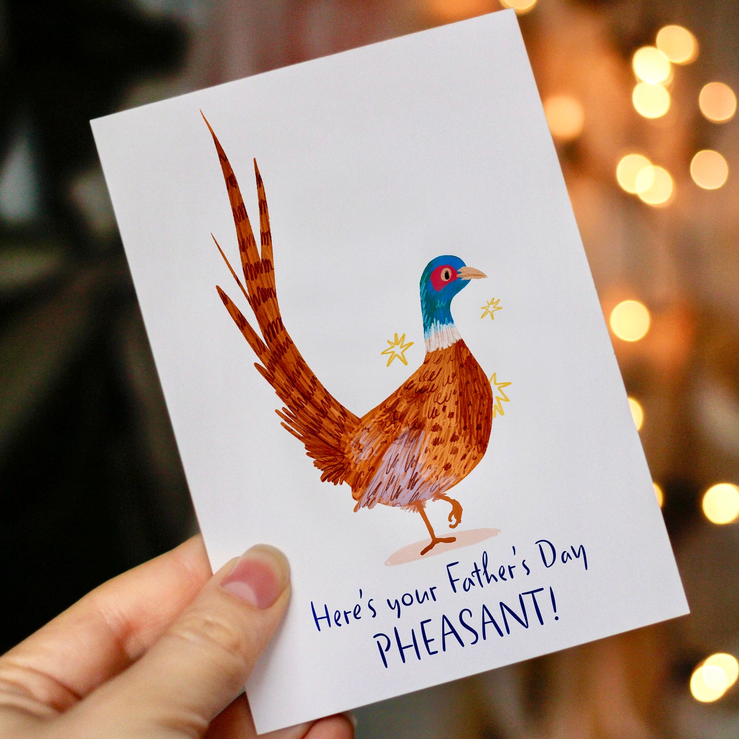 Card with a pheasant illustration and text 'Here's your Father's Day PHEASANT!' held by a hand with a blurred lights background.