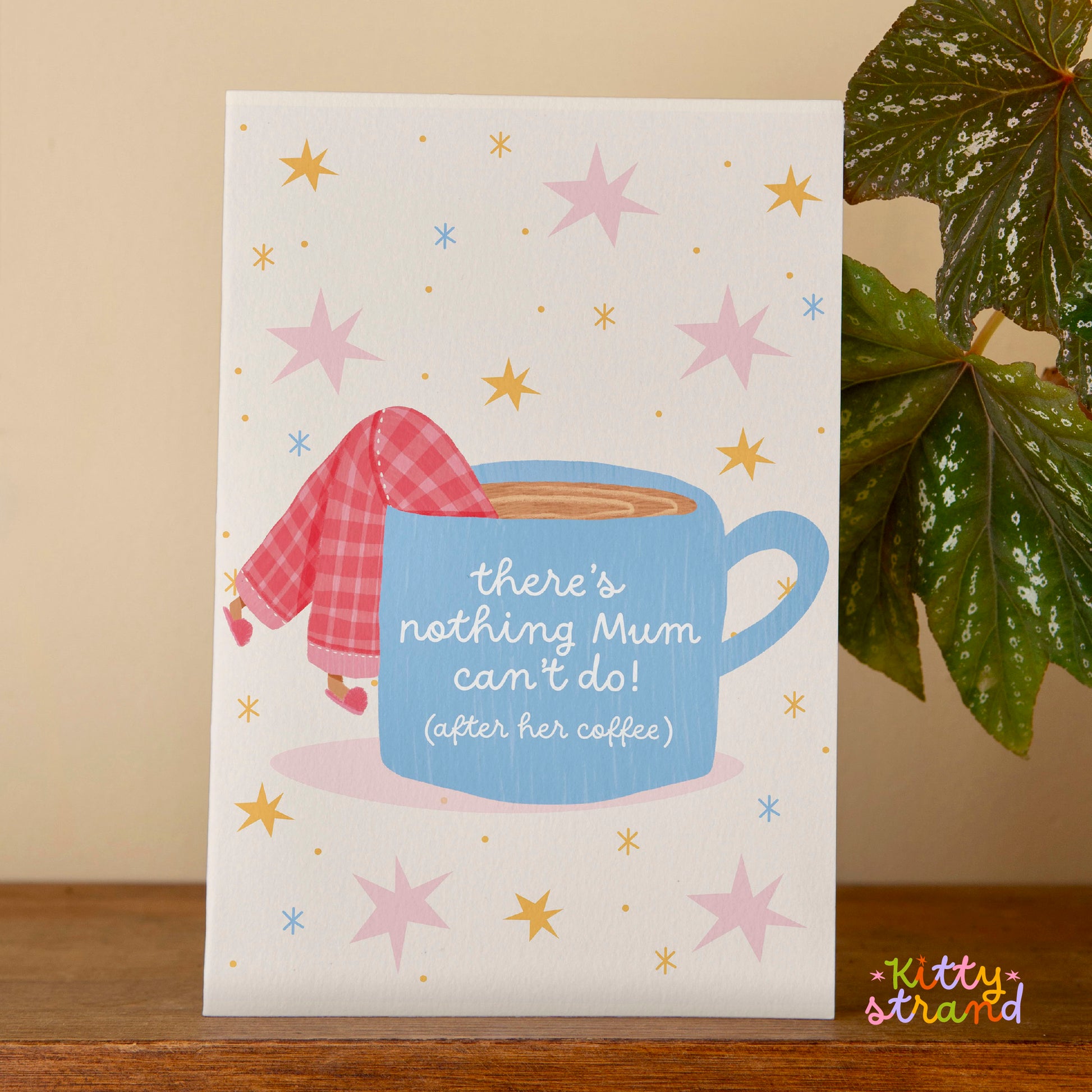 Card with a blue coffee cup and checkered shirt design, featuring stars and text, on a wooden surface with a plant in the background.