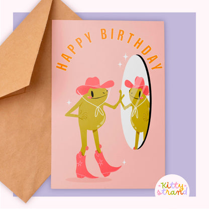 Birthday card with frog illustration wearing a hat and boots, accompanied by a brown envelope on a purple background.