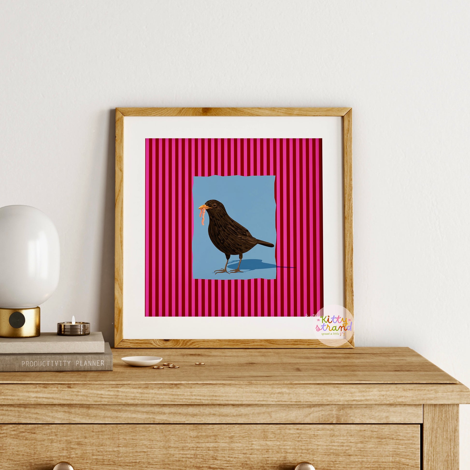 pink and blue bird-inspired wall art. Wall art with a vibrant striped pink background and a soft blue geometric square sits behind an original hand illustration of a blackbird with a worm in its mouth. Colourful, bold dopamine decor art print for nature lovers displayed in a wood frame