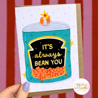 Card with a can of beans illustration and 'It's always bean you' text, held against a striped background.