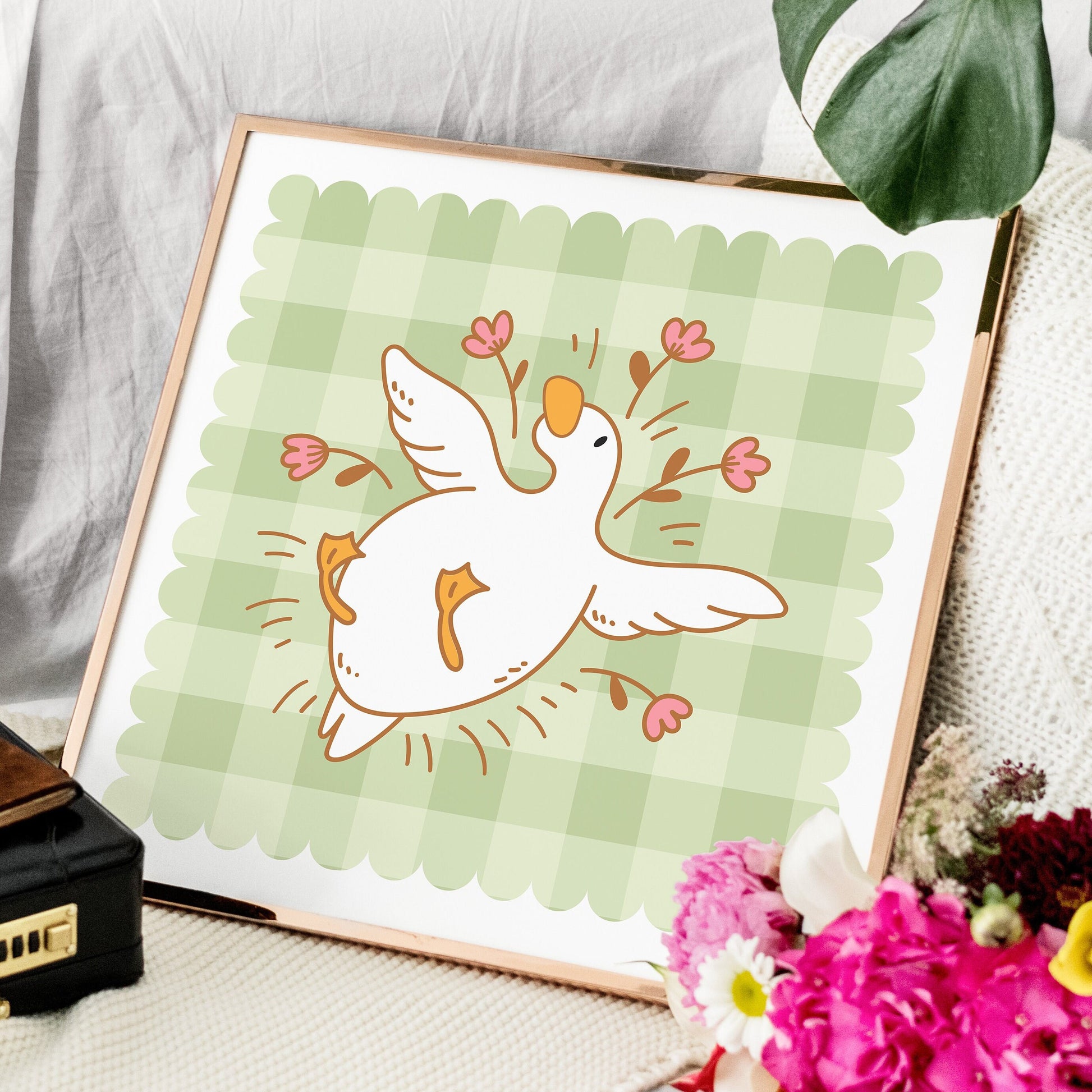 Framed illustration of a bird with flowers on a green checkered background, placed on a surface with flowers and a suitcase.