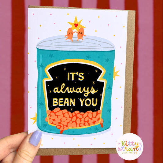 Card with a can of beans illustration and 'It's always bean you' text, held against a striped background.