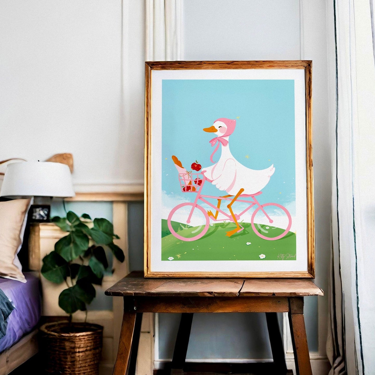 Framed artwork of a duck riding a bicycle with a pink basket resting on a bedside table 