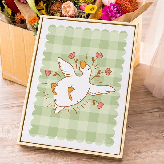 Framed illustration of a duck with flowers on a green checkered background, placed on a wooden surface with flowers in the background.